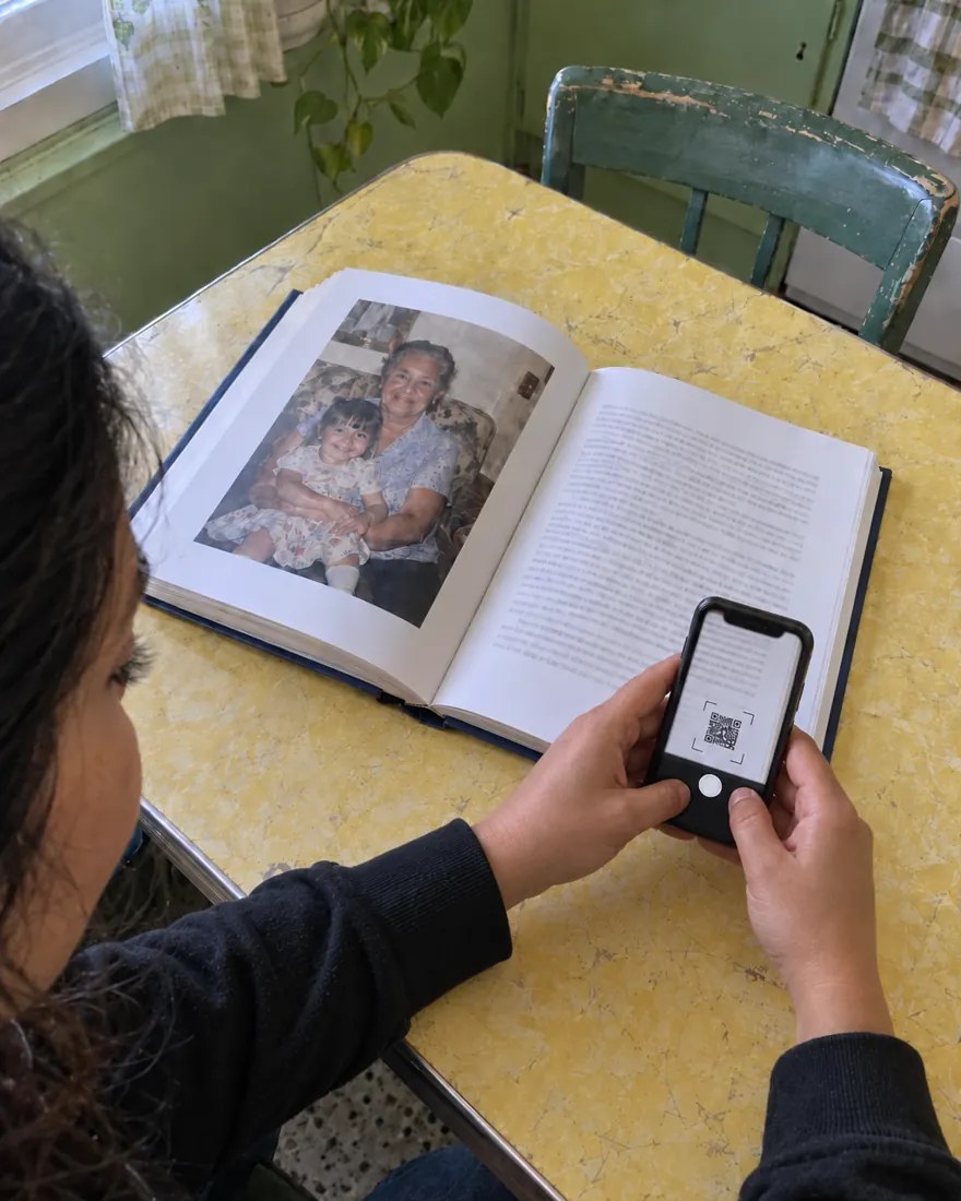 At the kitchen table, a granddaughter scans the printed code on a chapter of an open Fablr hardcover with her phone camera.