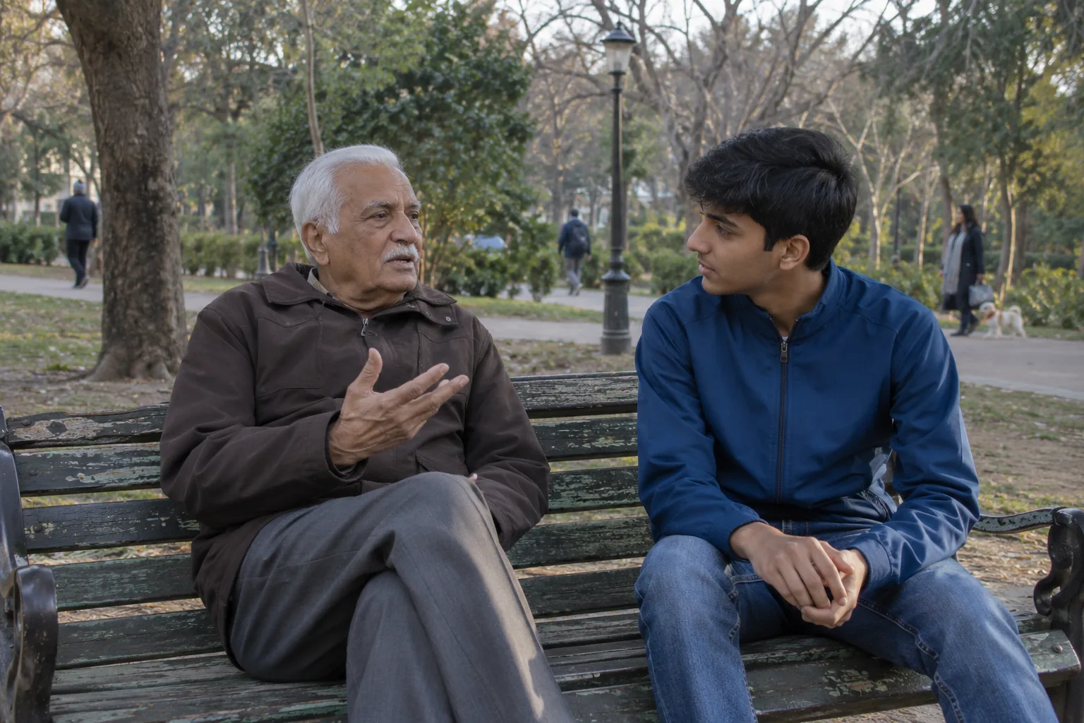 A grandfather on a park bench raises a hand mid-sentence as his teenage grandson listens.