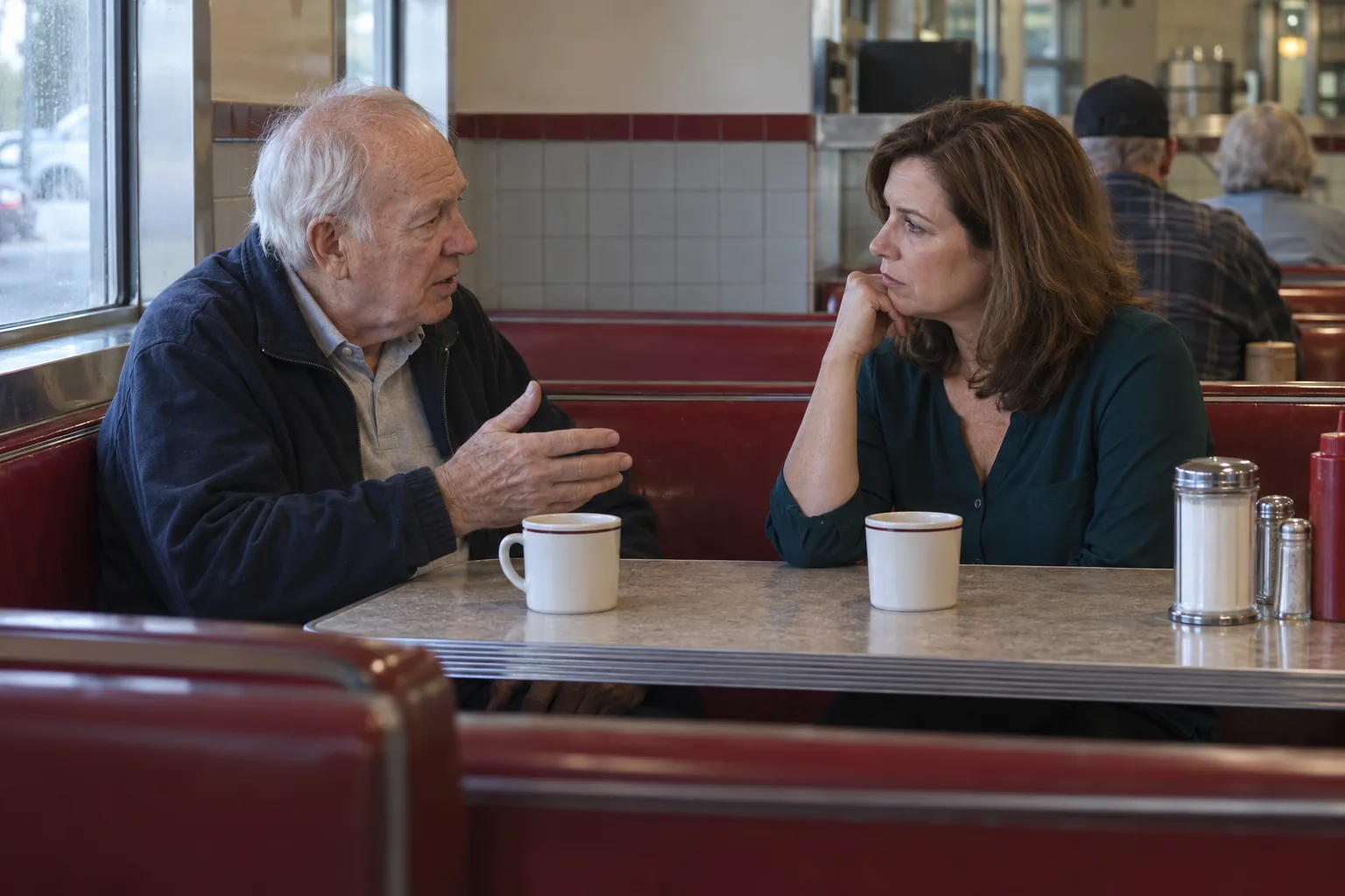 A father talks quietly with his adult daughter across a diner booth, coffee cups between them.